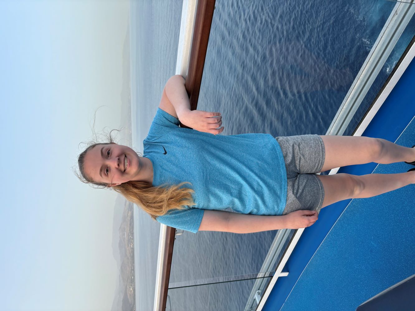 Grace smiles as she stands on the Odyssey, a cruise ship that she went on last summer with her family. The hazy skyline and ocean are in the background. She is wearing a blue t-shirt and gray shorts.